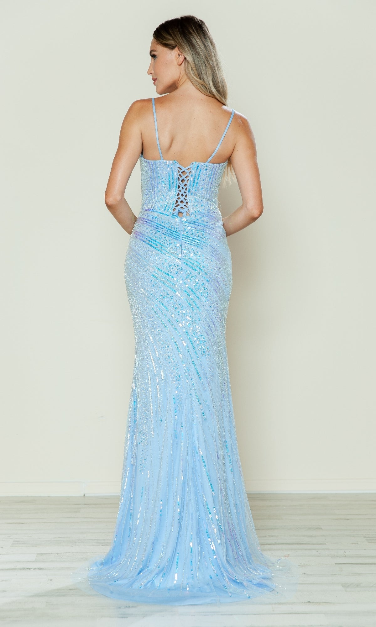 Woman wearing the Lace-Up Long Beaded Prom Dress: Poly USA 9438—a light blue, floor-length gown with thin straps and a lace-up back—stands on a light wood floor, facing away from the camera.
